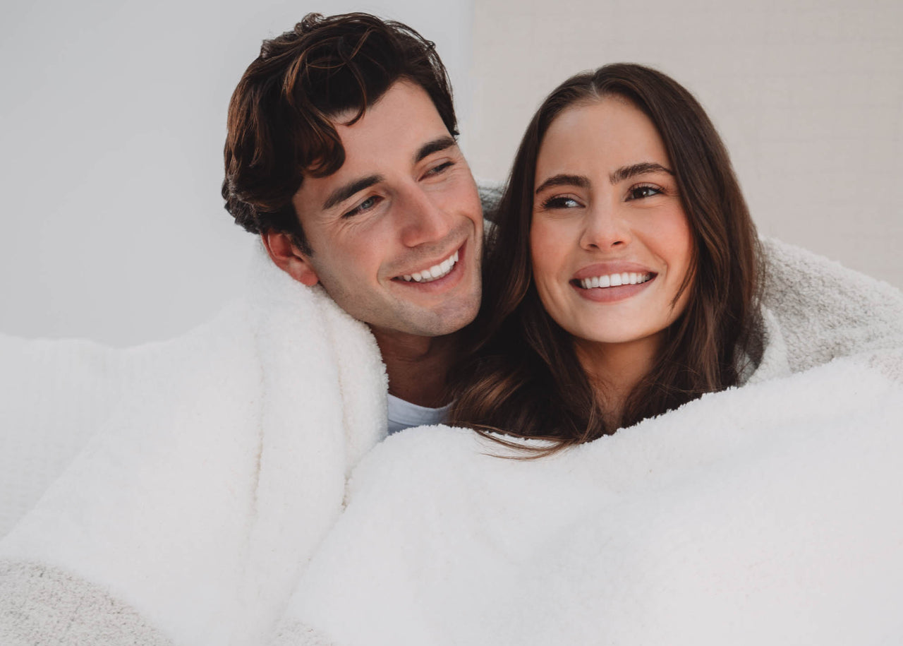 Man and woman smiling under a kashwere blanket, embracing each other.